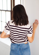 A woman viewed from behind wearing a short-sleeve knit top with dark brown and off-white horizontal stripes, paired with blue jeans.