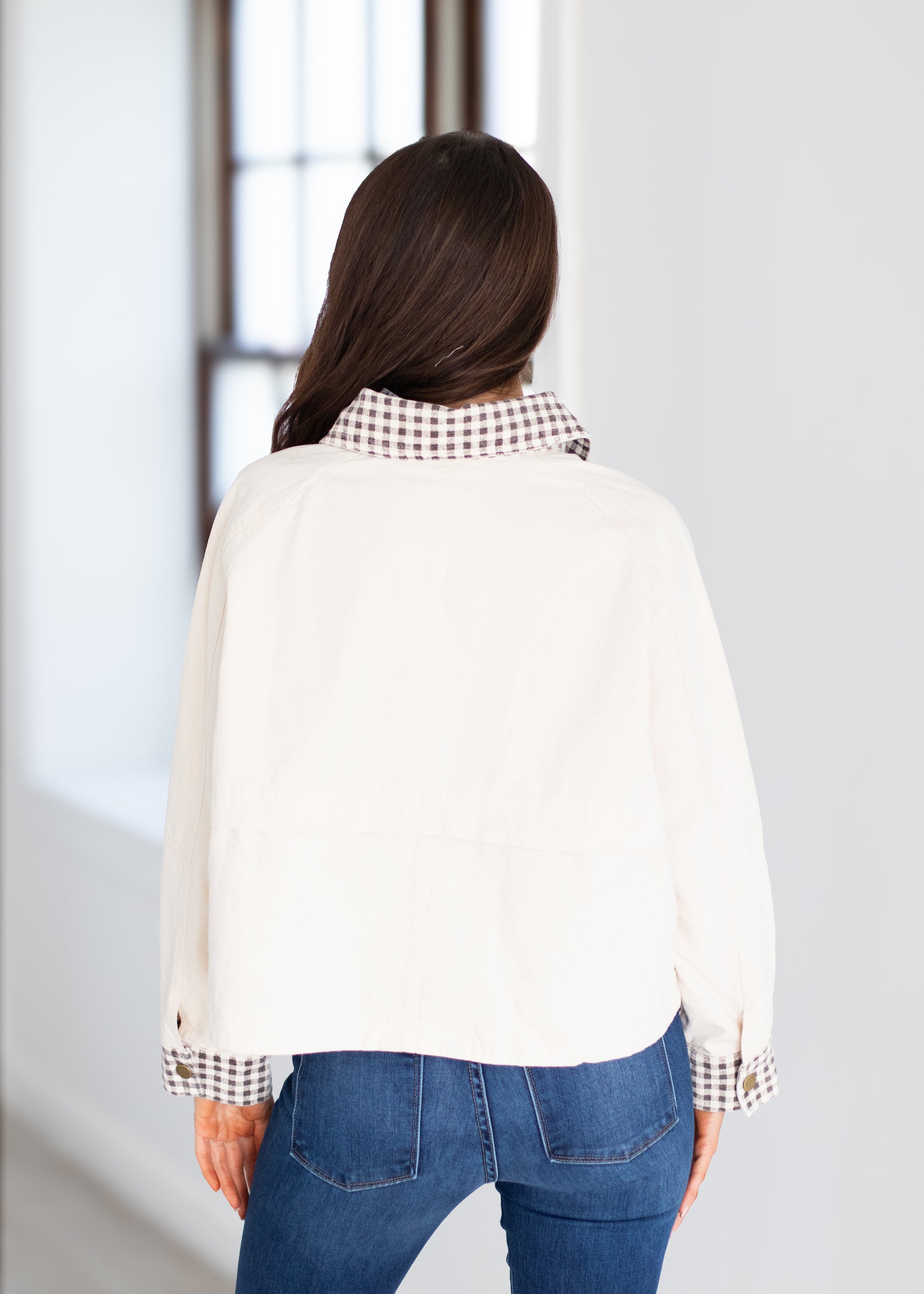 A woman viewed from behind wearing a cream-colored long-sleeve blouse with a brown and white checkered patterned collar and cuffs, paired with blue jeans.