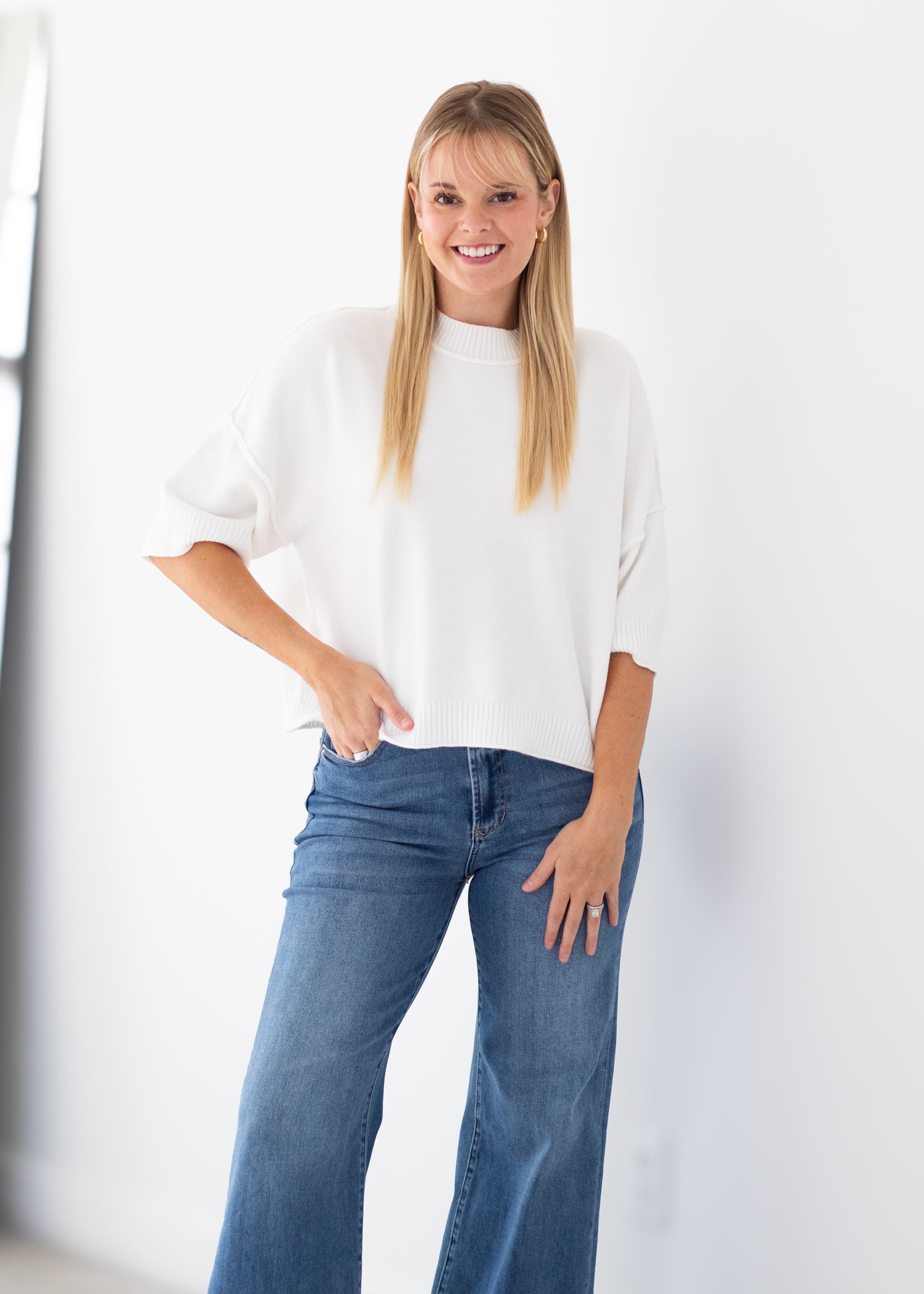 A woman modeling a white short-sleeve knit top with blue jeans, standing against a bright white wall.