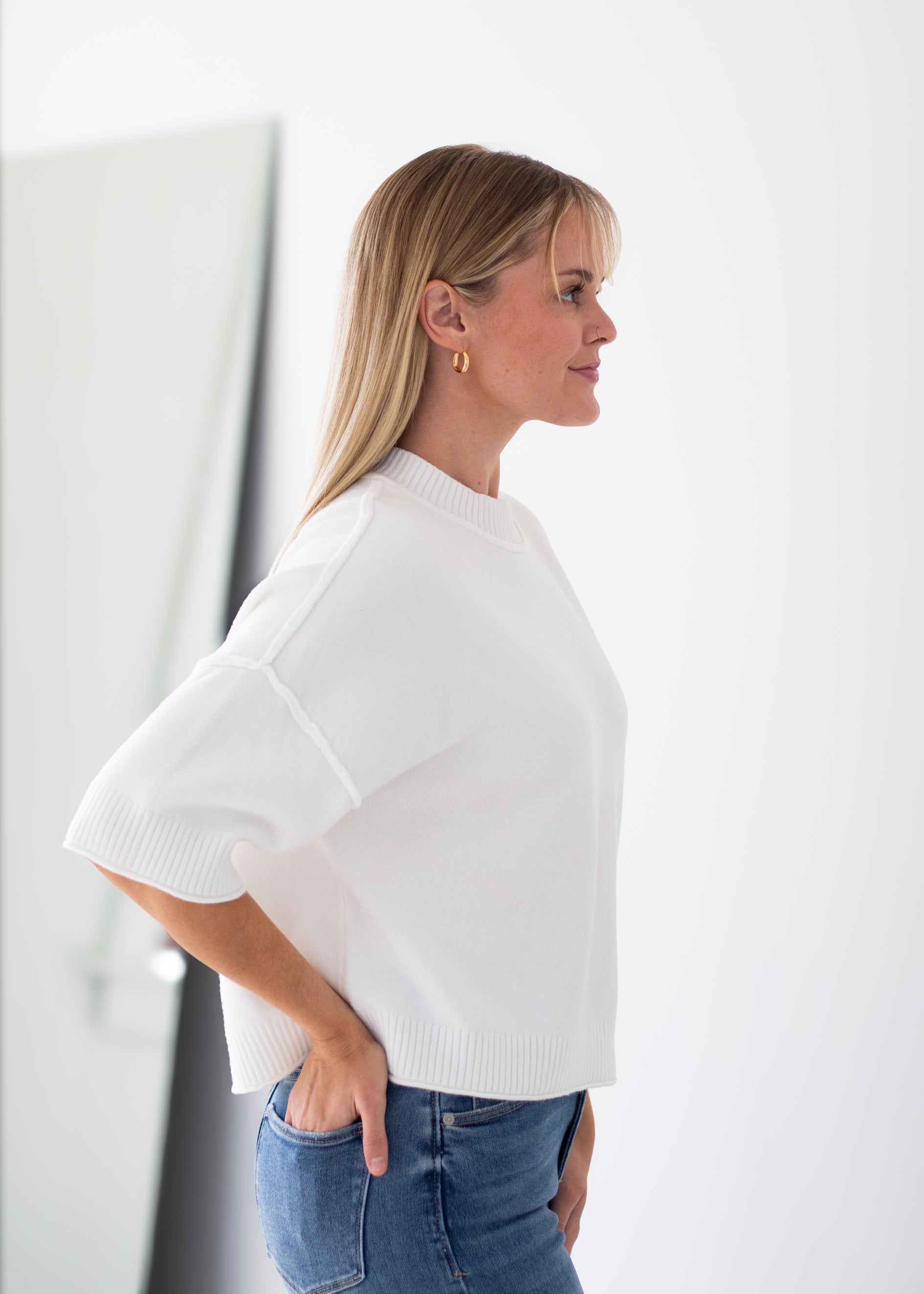 A woman in profile wearing a white short-sleeve knit top and blue jeans, standing against a bright white background.