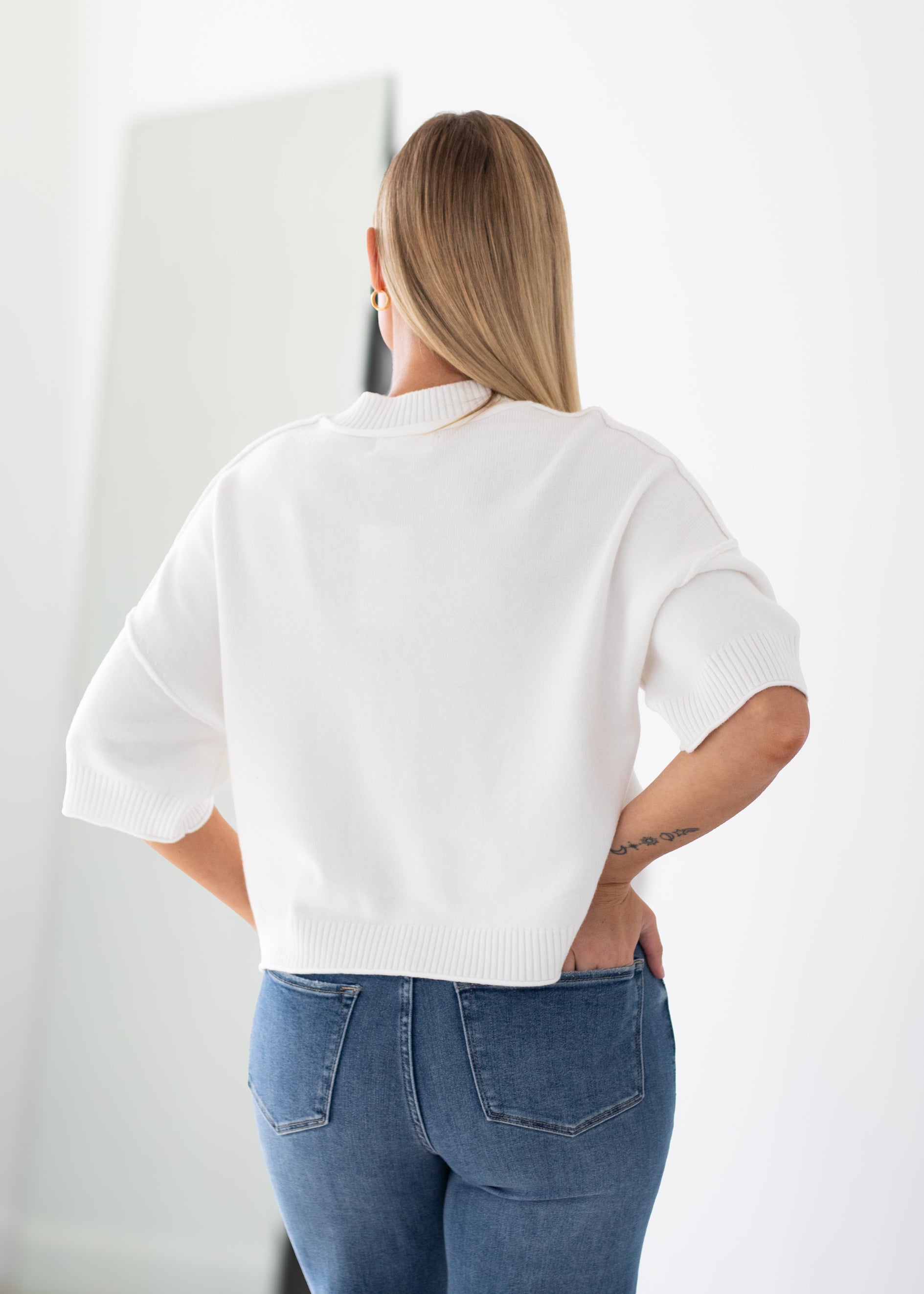 A woman viewed from behind wearing a white short-sleeve knit sweater and blue jeans in a bright indoor setting.