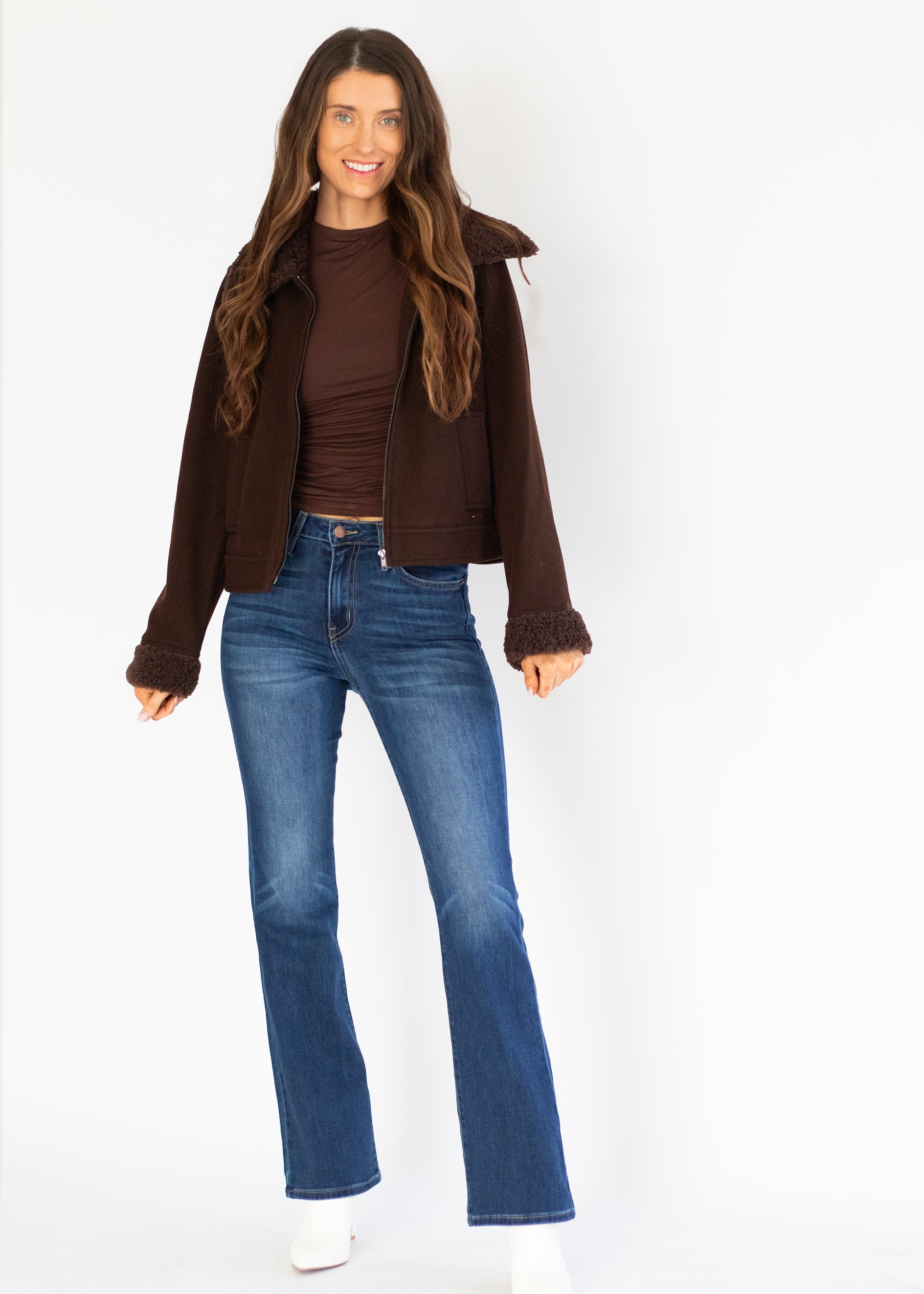 A woman modeling a dark brown cropped jacket with fuzzy cuffs, worn over a matching brown ribbed top and blue jeans, against a plain white studio background.