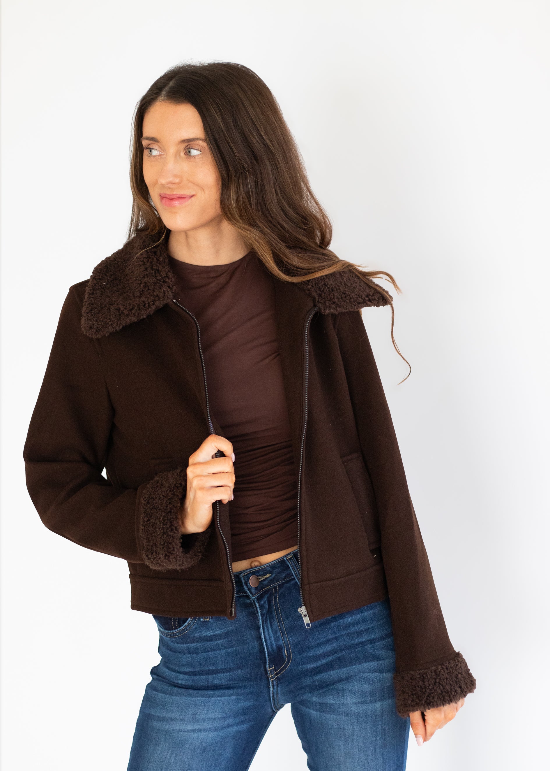 A woman wearing a dark brown suede-like jacket with a fuzzy shearling-style collar and cuffs, layered over a matching dark brown top and blue jeans, photographed against a plain white studio background.
