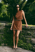 A woman wearing a brown matching set with a high neck and decorative chain-like trim, standing outdoors against a mossy stone wall and trees.