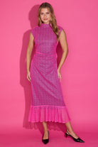 A woman modeling a bright pink sleeveless midi dress with a textured lace pattern, high mock neck, and ruffled hem, standing against a solid pink studio background.