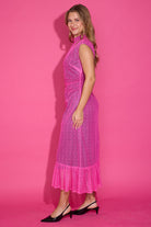 A woman modeling a bright pink sleeveless lace midi dress with a ruffled hem, standing against a solid pink studio backdrop.