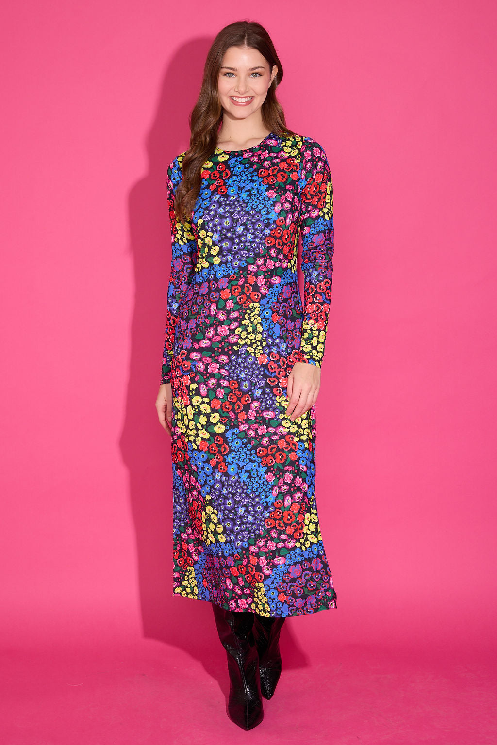 A woman modeling a long-sleeve midi dress with a vibrant multicolor floral pattern, standing against a solid pink studio background.