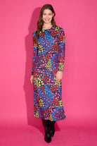 A woman modeling a long-sleeve midi dress with a vibrant multicolor floral pattern, standing against a solid pink studio background.