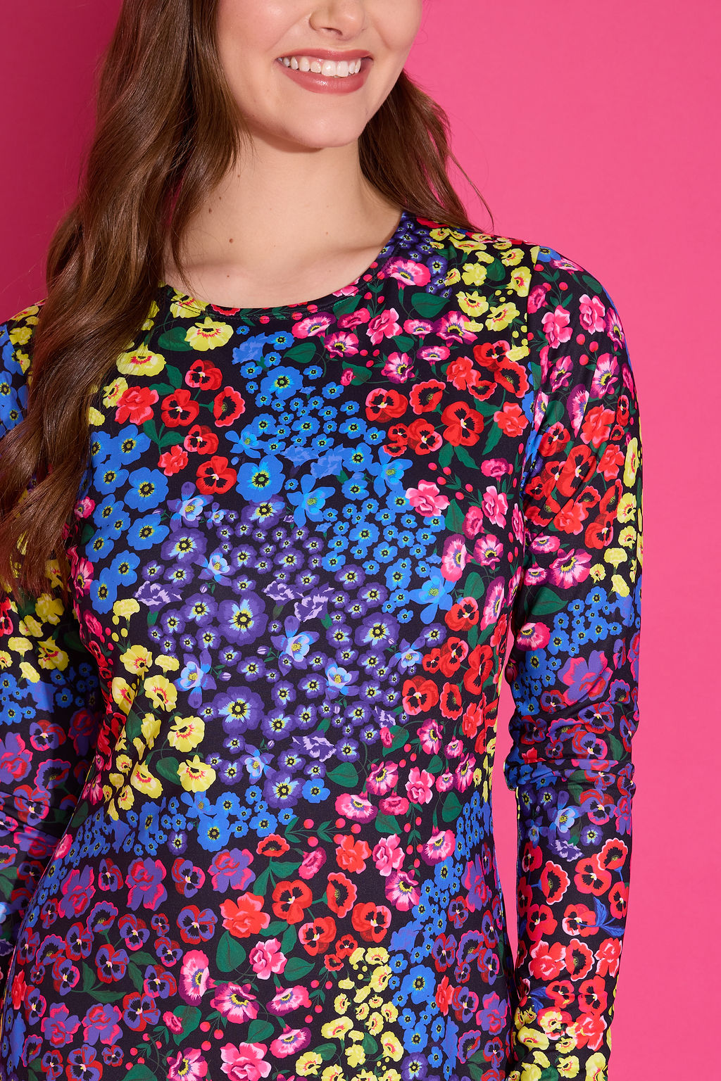 A woman wearing a long-sleeve crewneck top with a vibrant multicolor floral pattern, set against a solid bright pink background.