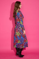 A woman wearing a long-sleeve midi dress with a vibrant multicolor floral pattern, paired with black knee-high boots, standing against a solid pink studio backdrop.