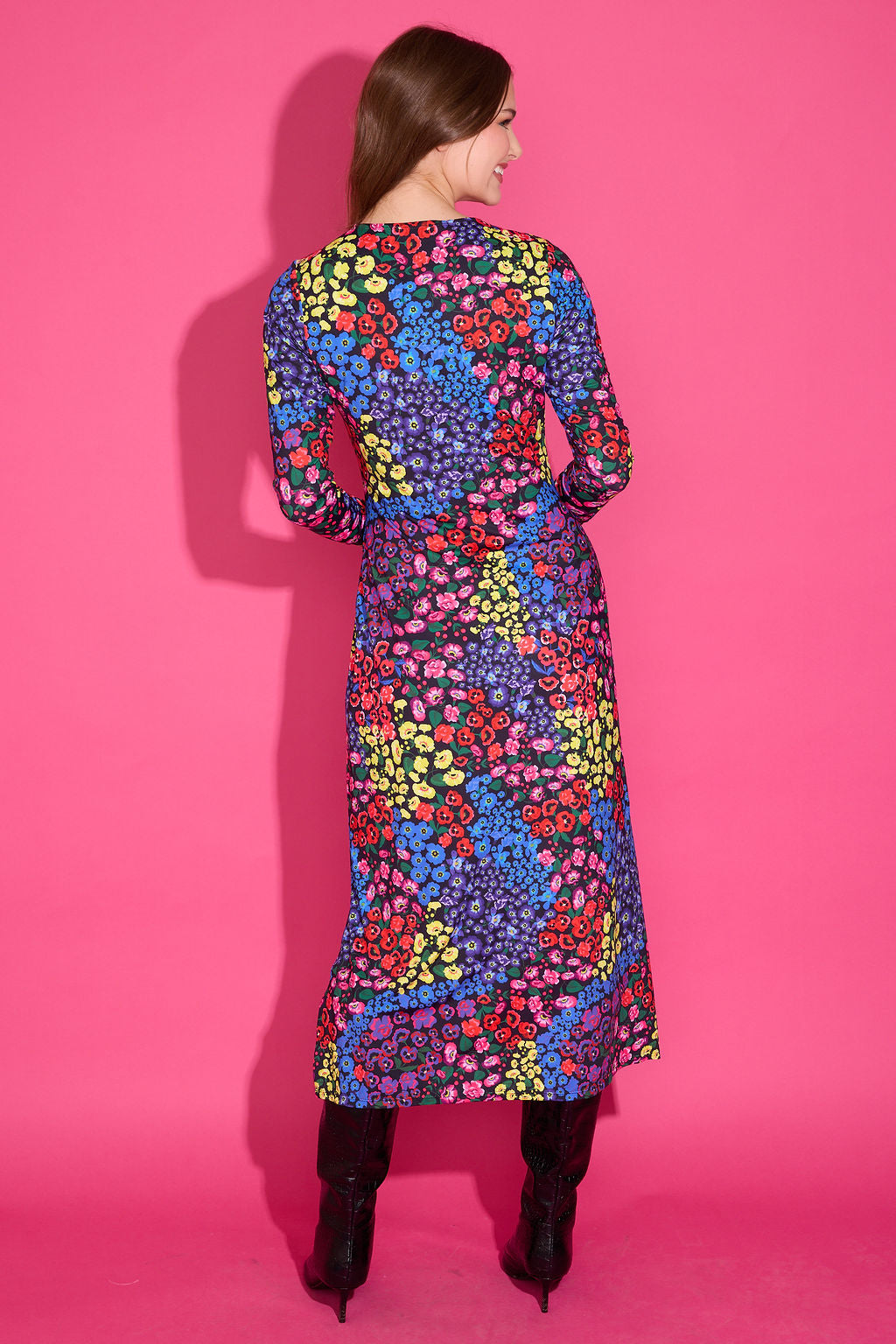 A woman viewed from the back wearing a long-sleeve midi dress with a vibrant multicolor floral pattern, paired with black ankle boots, set against a solid pink studio background.
