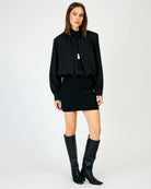 A model stands against a light gray studio background wearing a black long-sleeve blouse with a drawstring neck, a black mini skirt, and black knee-high boots.
