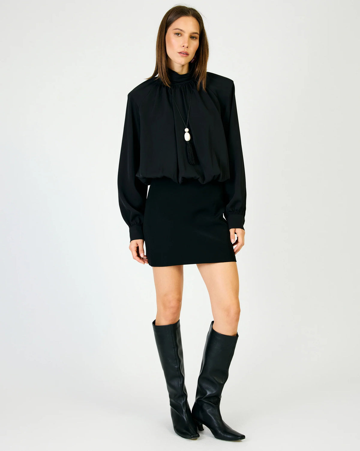 A model stands against a light gray studio background wearing a black long-sleeve blouse with a drawstring neck, a black mini skirt, and black knee-high boots.