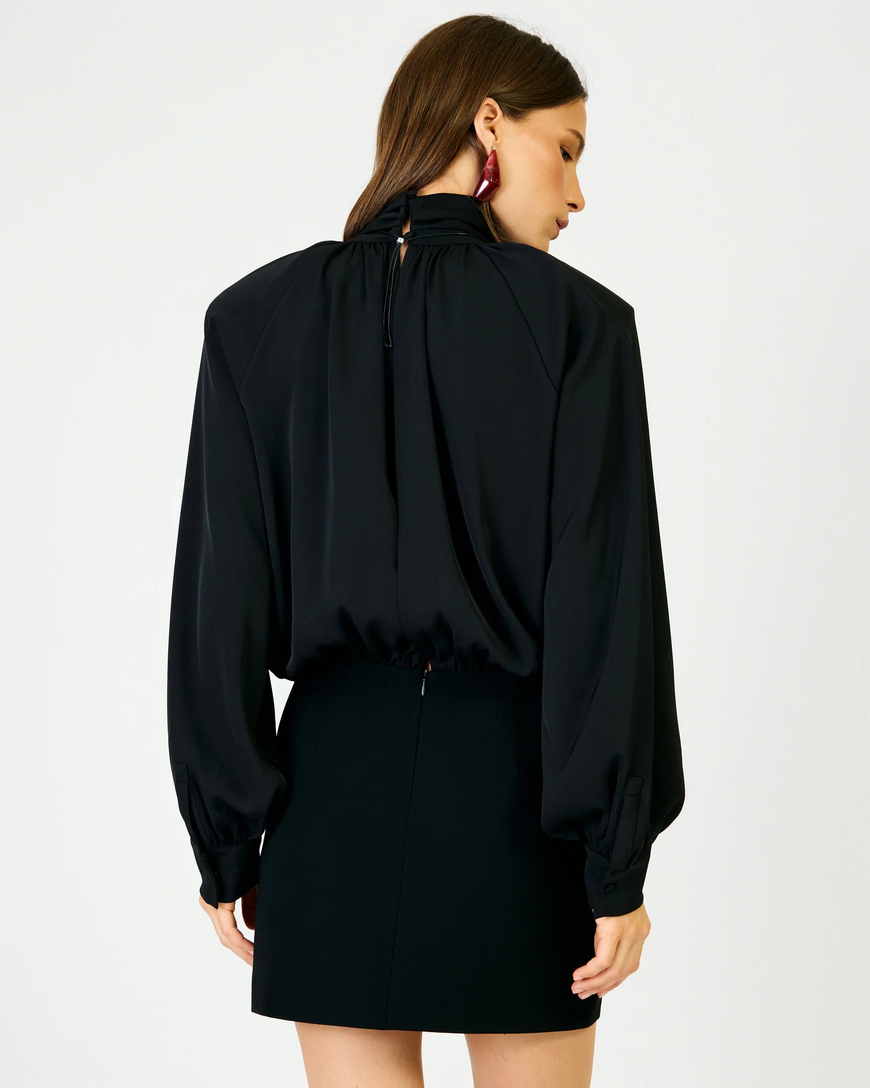 A woman shown from the back wearing a black long-sleeve blouse with a high gathered neckline and a black mini skirt against a plain light gray studio background.