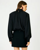 A woman shown from the back wearing a black long-sleeve blouse with a high gathered neckline and a black mini skirt against a plain light gray studio background.