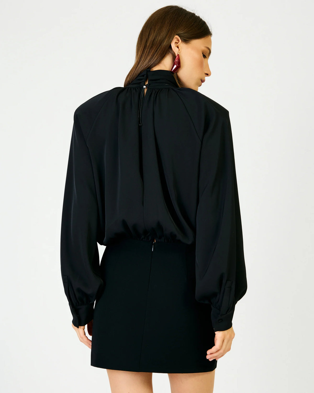 A woman shown from the back wearing a black long-sleeve blouse with a high gathered neckline and a black mini skirt against a plain light gray studio background.