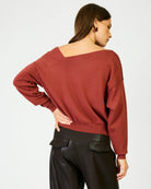 A woman shown from the back wearing a rust-colored, off-the-shoulder knit sweater and black pants against a plain white background.