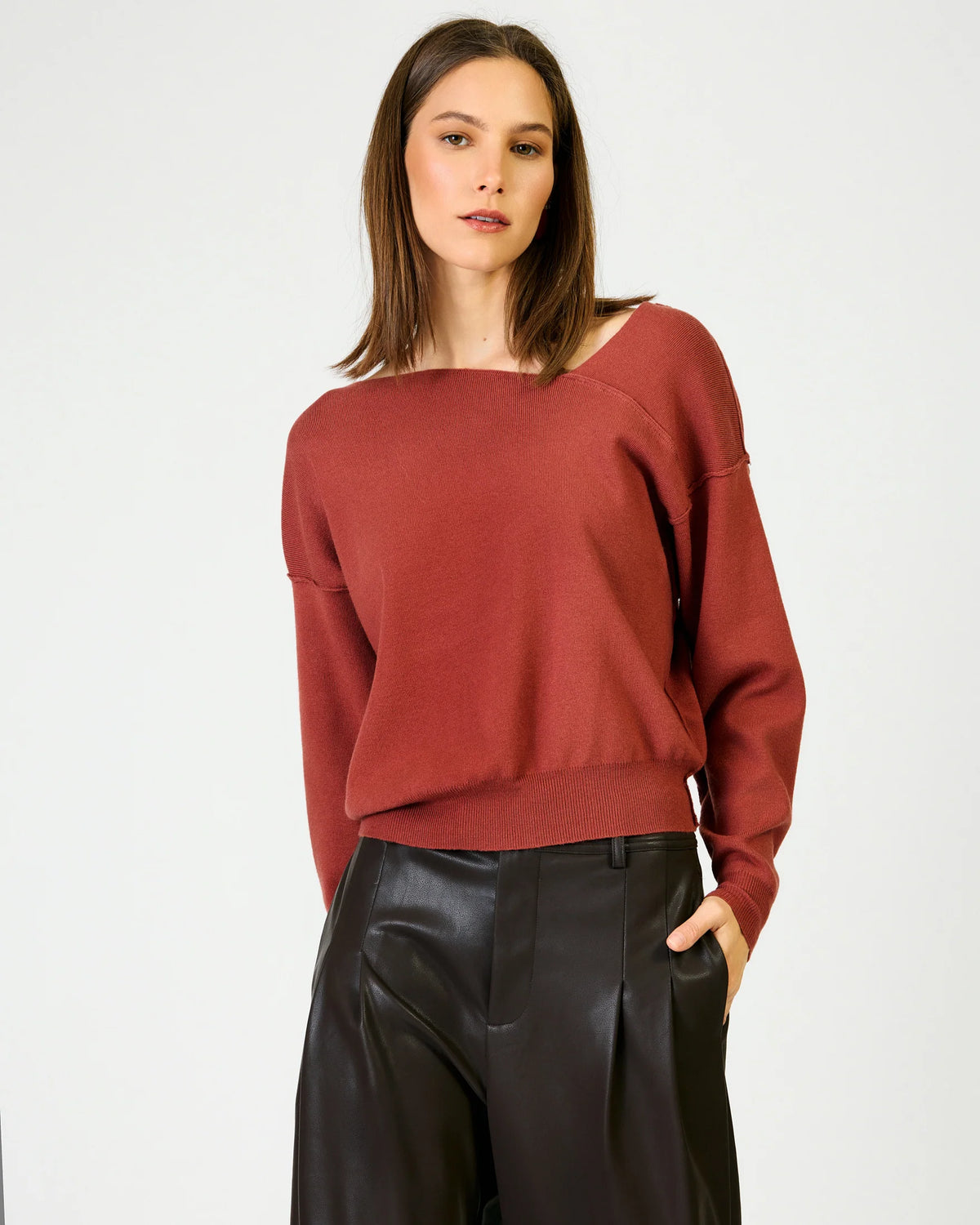 A woman wearing a rust-red knit sweater with a wide boat neckline and black leather pants, standing against a plain white studio background.