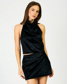 A woman modeling a black two-piece set consisting of a halter crop top and a matching mini skirt, photographed against a plain light gray studio background.