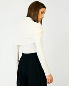 A woman shown from the back wearing a white draped long-sleeve top and black pleated trousers, photographed against a clean white studio background.
