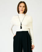 A woman modeling a cream draped mock-neck top with long sleeves, paired with black pleated trousers and a long black tassel necklace, against a plain light gray background.