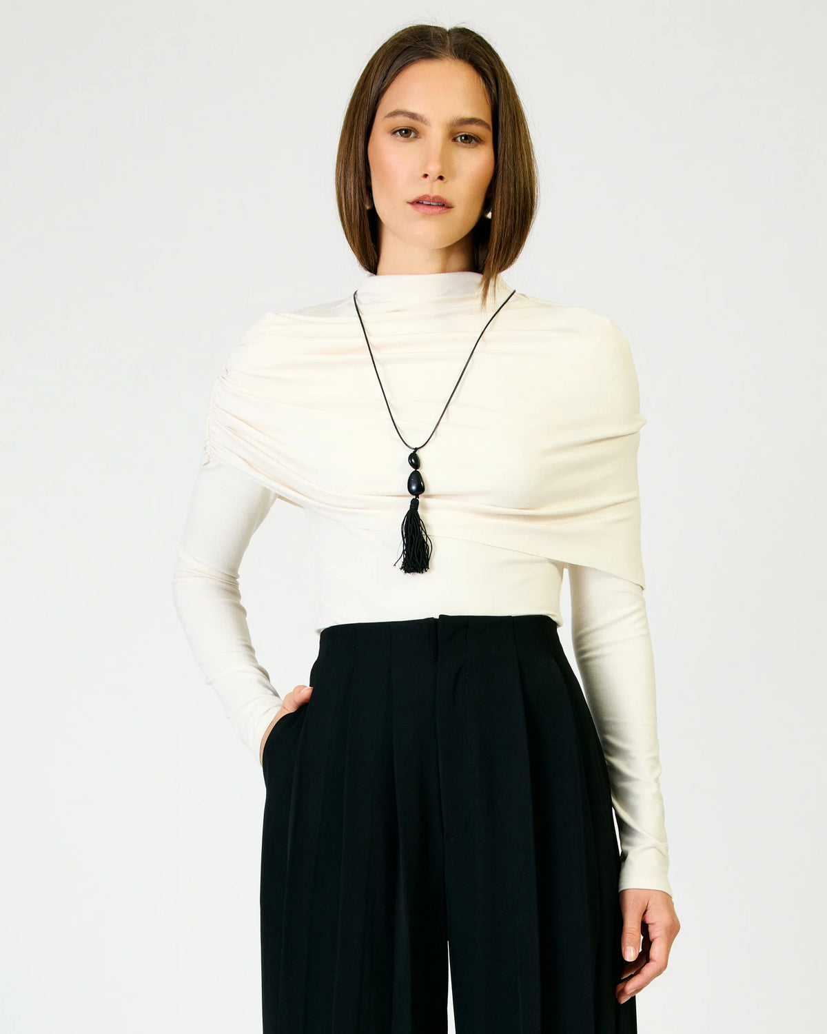A woman modeling a cream draped mock-neck top with long sleeves, paired with black pleated trousers and a long black tassel necklace, against a plain light gray background.