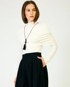 A woman with a shoulder-length brown bob model is wearing a cream-colored draped long-sleeve top, black high-waisted pleated trousers, and a long black tassel necklace. She is photographed against a clean, light off-white studio background.