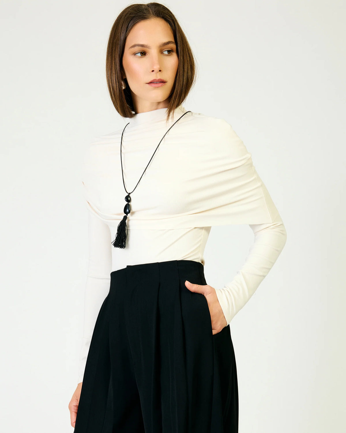 A woman with a shoulder-length brown bob model is wearing a cream-colored draped long-sleeve top, black high-waisted pleated trousers, and a long black tassel necklace. She is photographed against a clean, light off-white studio background.