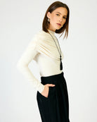 A woman modeling a cream long-sleeve mock-neck top with gathered draping details on the chest and black high-waisted trousers, set against a plain white studio background.