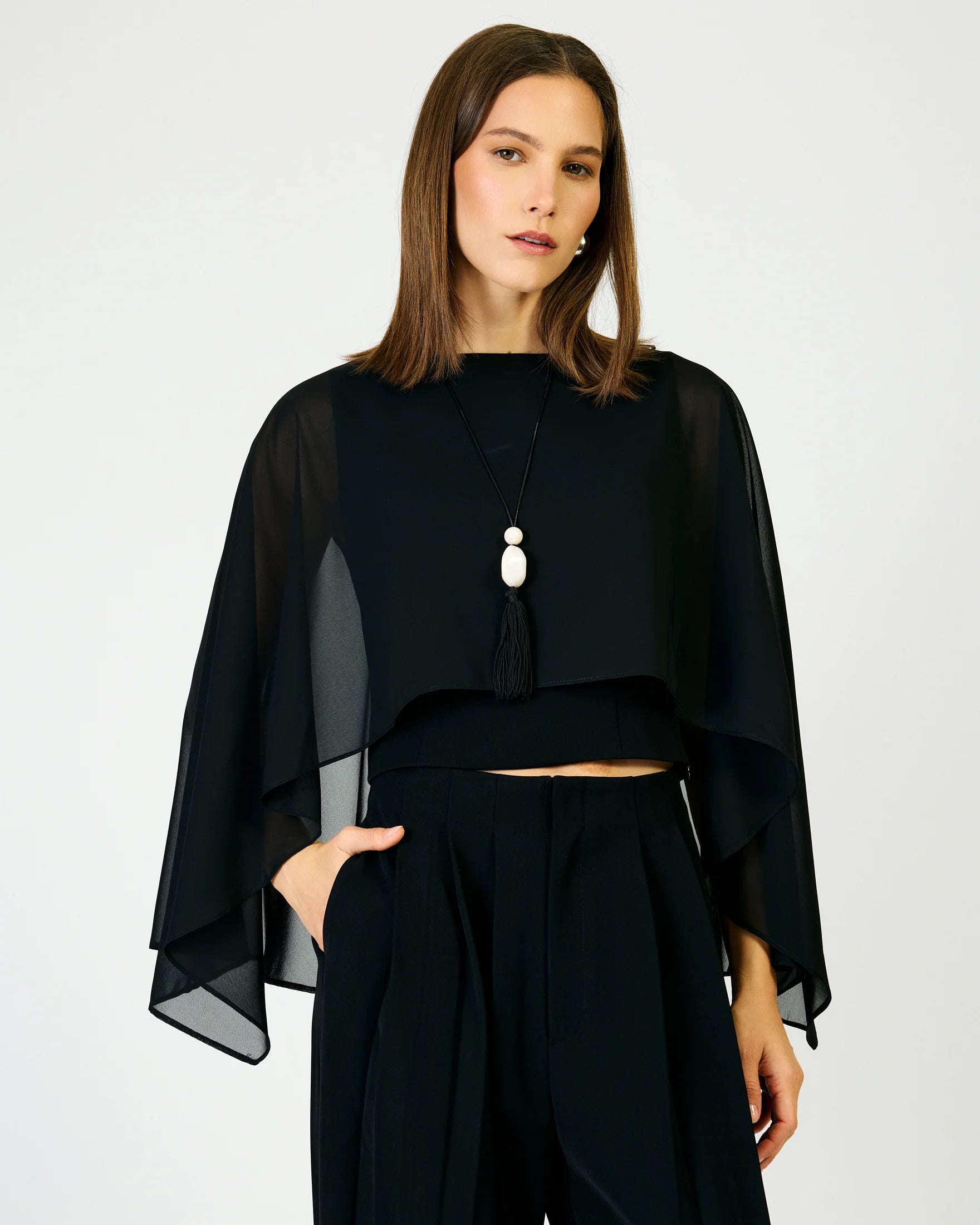 A woman modeling a black sheer draped cape-style top with wide bell sleeves, paired with black high-waisted trousers and a long pendant necklace, against a plain light gray studio background.