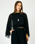 A woman modeling a black sheer draped cape-style top with wide bell sleeves, paired with black high-waisted trousers and a long pendant necklace, against a plain light gray studio background.
