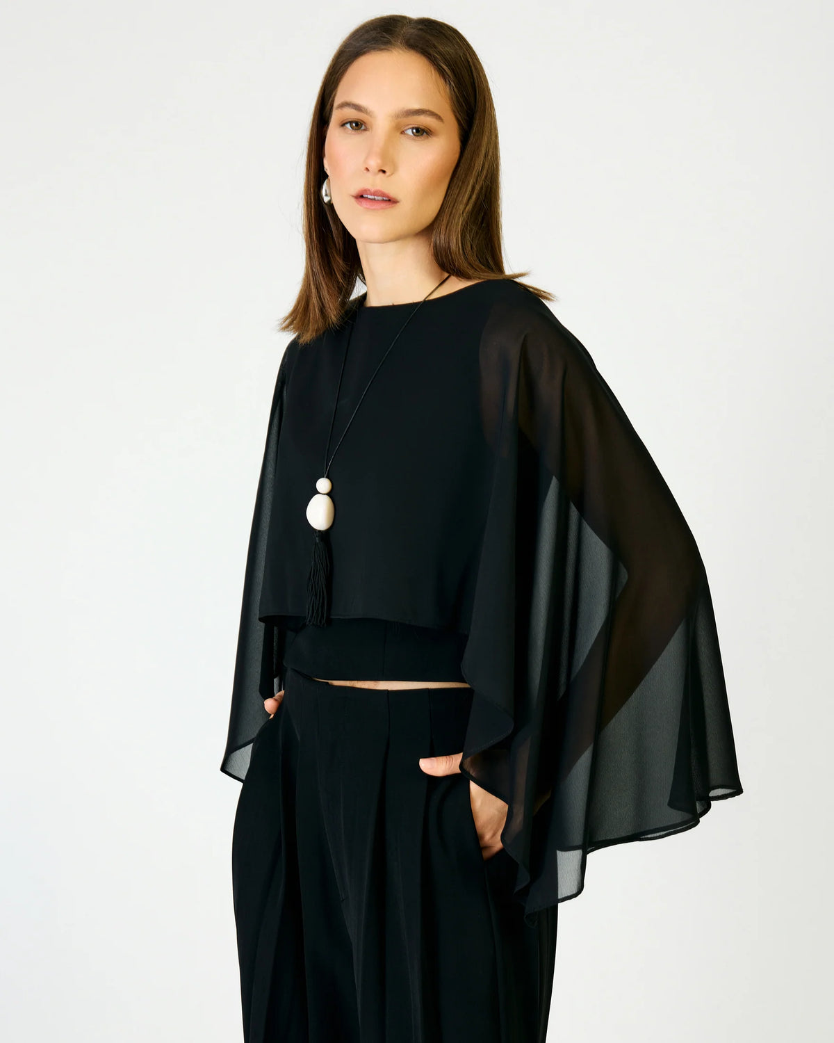A woman modeling a black sheer cape-style top with draped sleeves and matching black pants against a plain light gray studio background.