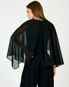 A woman shown from the back wearing a black sheer cape-style top over a black outfit, photographed against a plain light background.