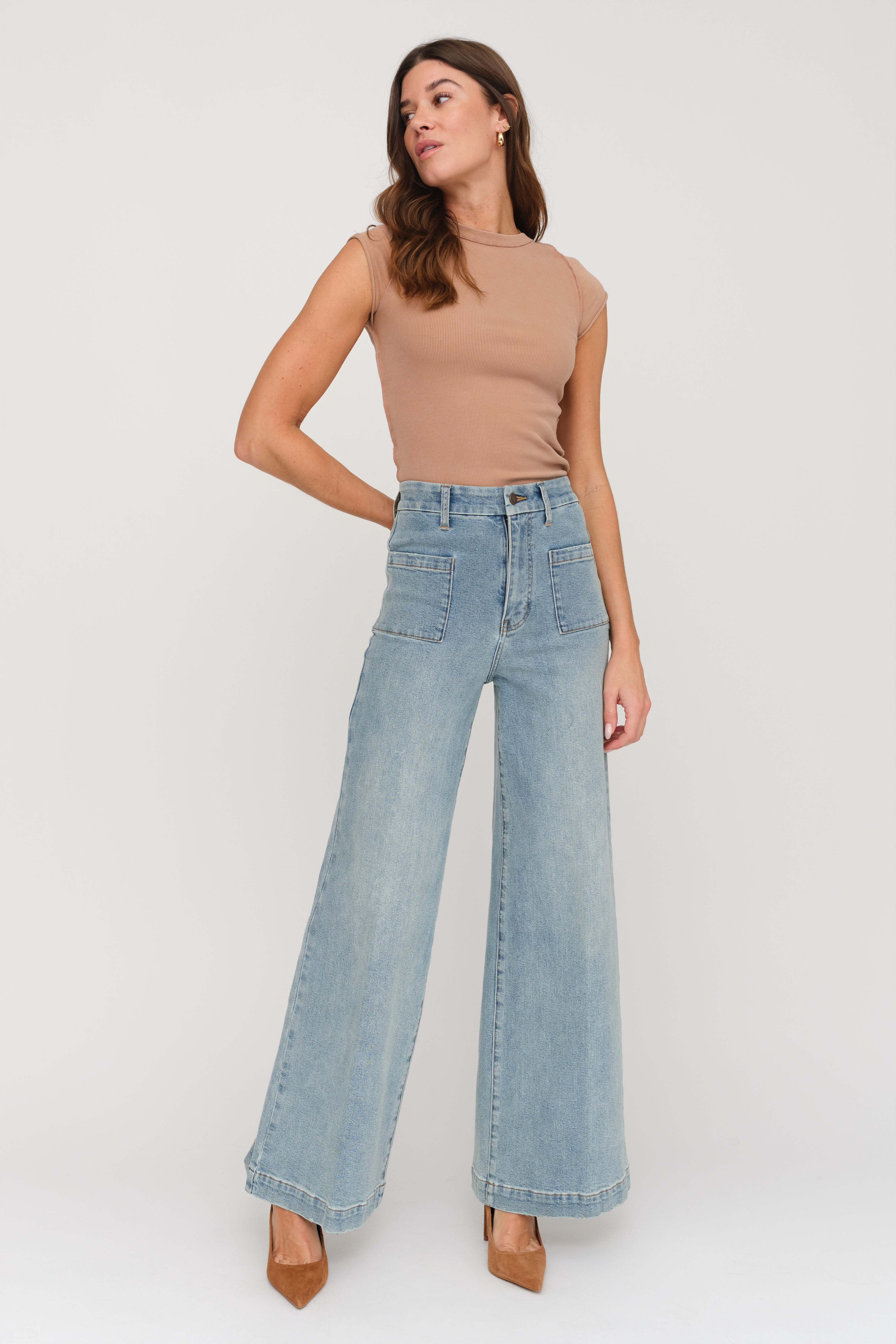 A woman modeling a pair of high-waisted, wide-leg light blue denim jeans with a tan sleeveless top, standing against a plain light gray studio background.