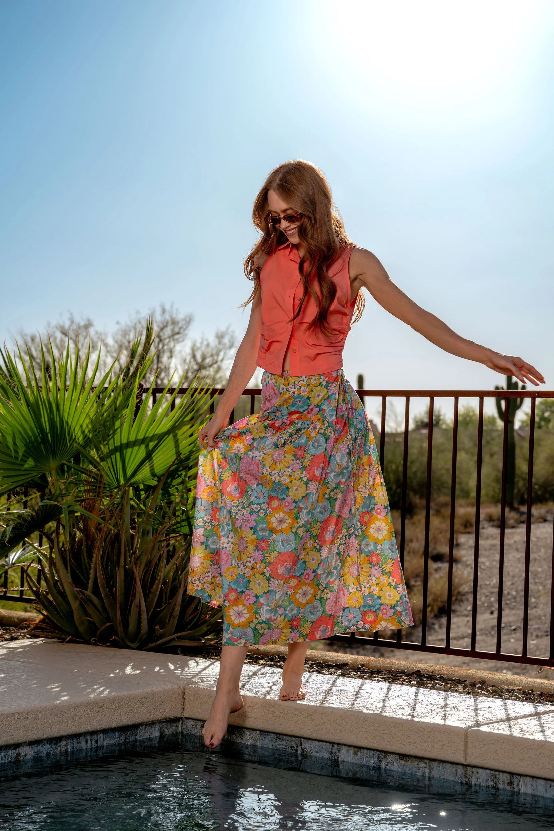 Woman in a colorful floral skirt and red top standing by a pool with a palm tree in the background.