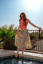 Woman in a colorful floral skirt and red top standing by a pool with a palm tree in the background.