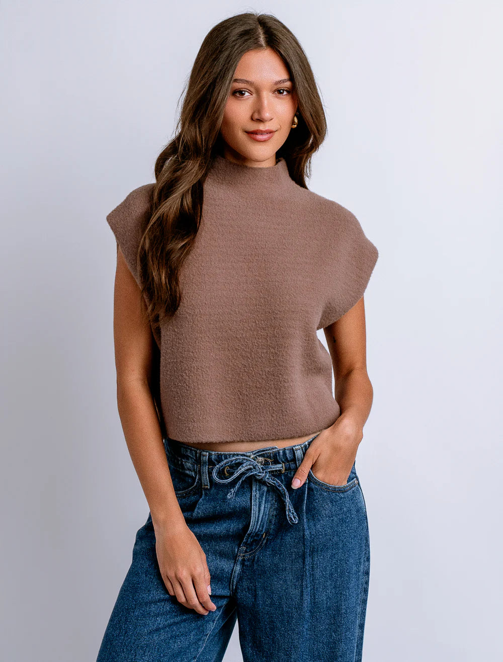 A woman wearing a cropped, short-sleeve knit top in a warm taupe-brown color, paired with blue jeans, against a plain light gray studio background.