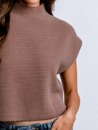 A close-up of a person wearing a fuzzy, short-sleeve mock-neck top in a dusty mauve-brown color, paired with blue jeans against a light gray background.