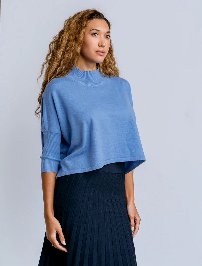 Woman wearing a blue sweater and dark pleated skirt against a light gray background