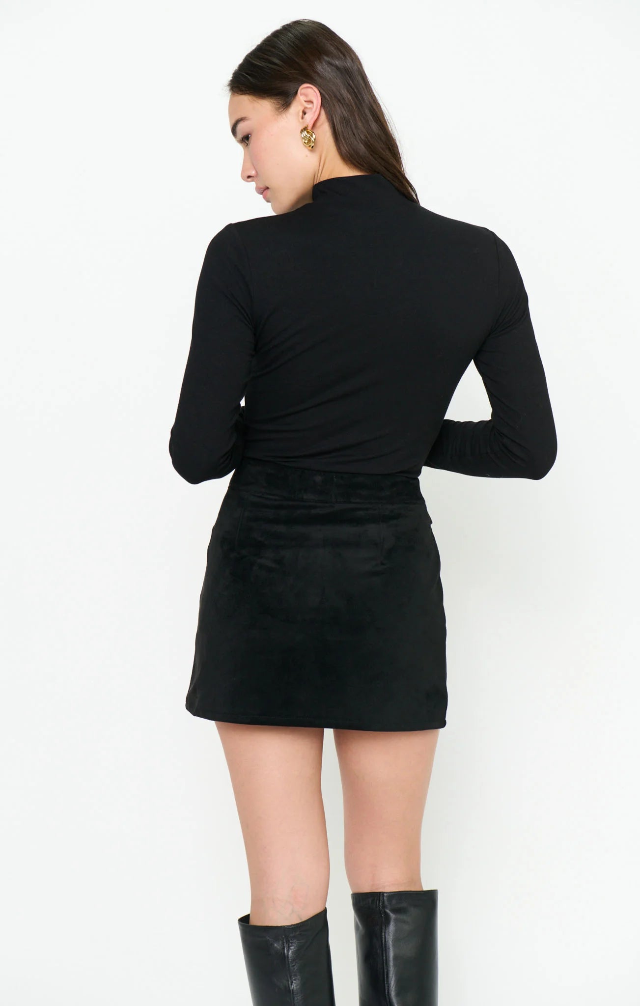 A woman shown from the back wearing a fitted black long-sleeve mock-neck top and a black mini skirt, paired with black knee-high boots, against a plain white studio background.
