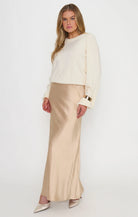 A woman modeling a cream long-sleeve knit top and a champagne beige satin maxi skirt dress, styled with pointed-toe heels against a plain white studio background.
