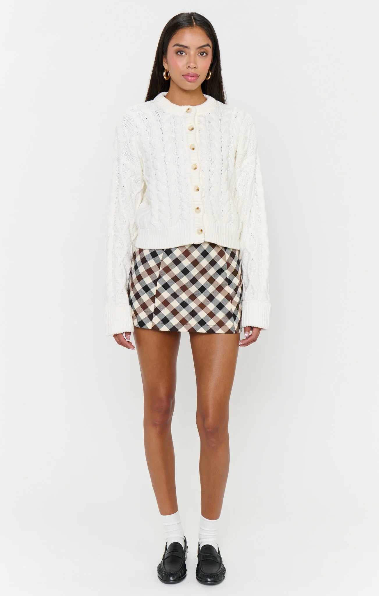 A model stands against a plain white studio background wearing a white cable-knit cropped cardigan with gold buttons, a brown, beige, and white plaid mini skirt, white socks, and black loafers.