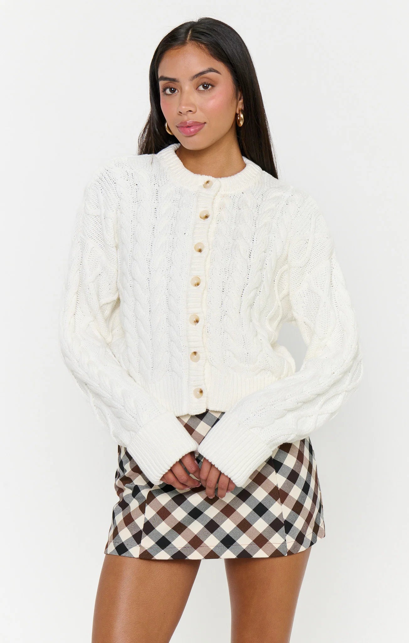 A woman modeling a cream cable-knit cropped button-up cardigan with a mock neck, paired with a brown, beige, and white gingham mini skirt, against a plain white studio background.