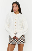 A woman modeling a cream cable-knit cropped button-up cardigan with a mock neck, paired with a brown, beige, and white gingham mini skirt, against a plain white studio background.