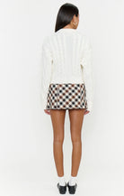 A woman shown from the back wearing a cream cable-knit cardigan, a brown, beige, and white plaid mini skirt, white socks, and black shoes, against a plain white studio background.