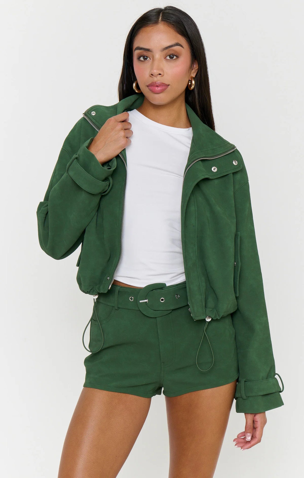 A woman modeling a dark green cropped jacket and matching belt-and-shorts set over a white top, photographed against a clean light gray studio background.