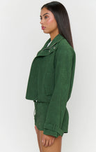 A woman shown in side profile wearing a dark green cropped jacket and matching green shorts against a plain light gray studio background.