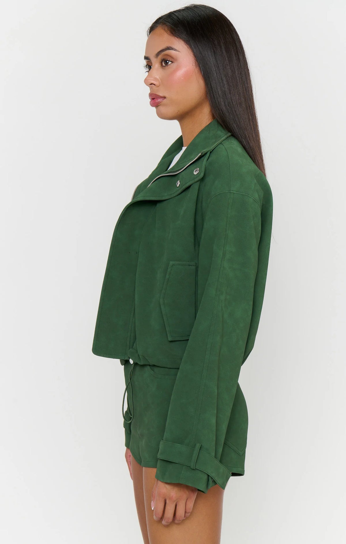 A woman shown in side profile wearing a dark green cropped jacket and matching green shorts against a plain light gray studio background.