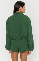 A woman shown from the back wearing a dark green long-sleeve collared top and matching shorts against a plain light gray studio background.