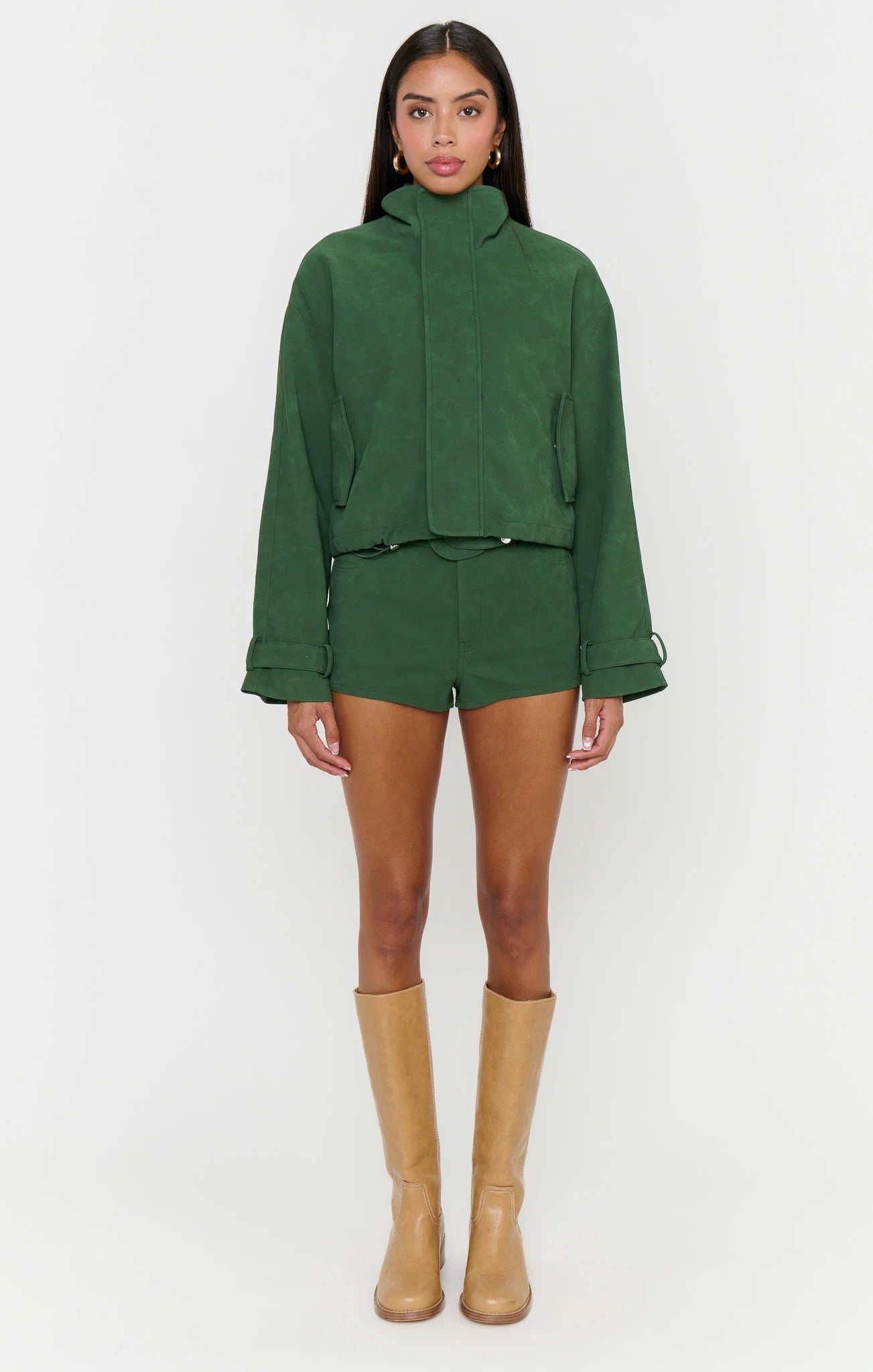 A model stands against a light gray studio background wearing a matching dark green cropped jacket and shorts set, tan knee-high boots, and gold hoop earrings.